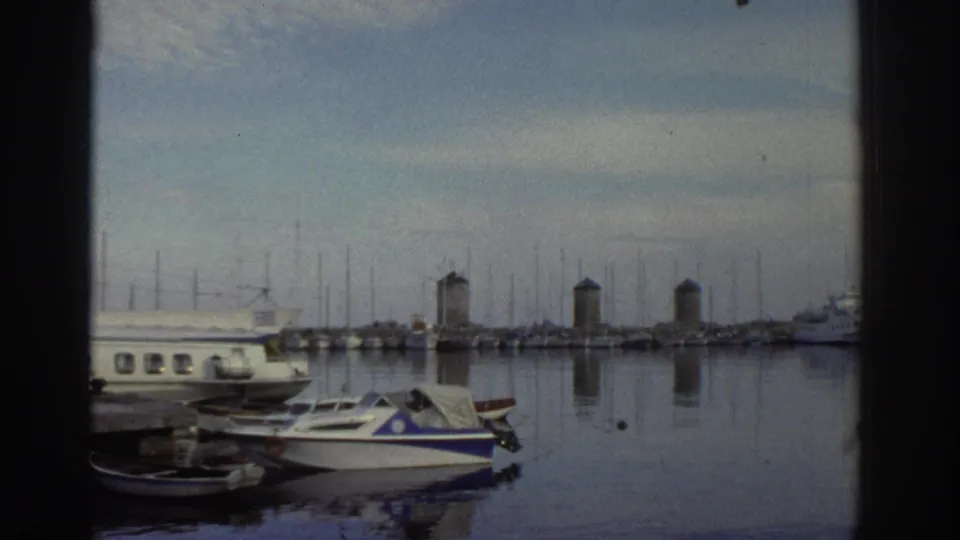 Greek harbour in 1976 — calm reflective water, moored motorboats and yachts, two background tower-like structures, no people visible.