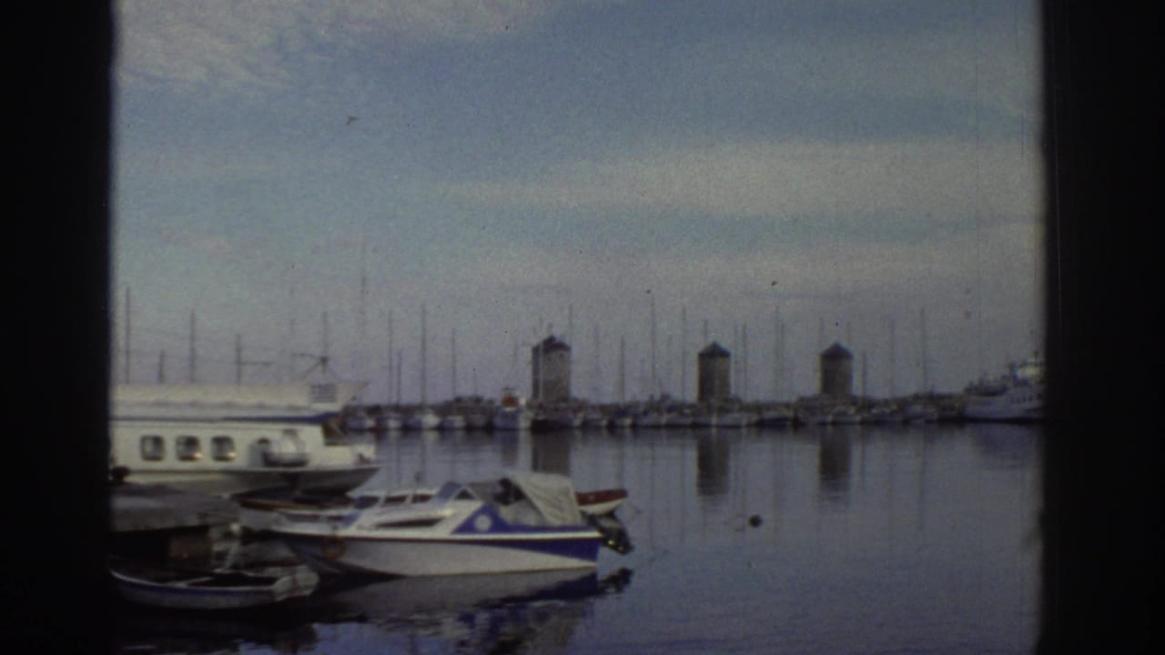 Greek harbor marina circa 1976; small motorboats moored at dock with stone windmill towers in middle distance, filmed on 8mm.