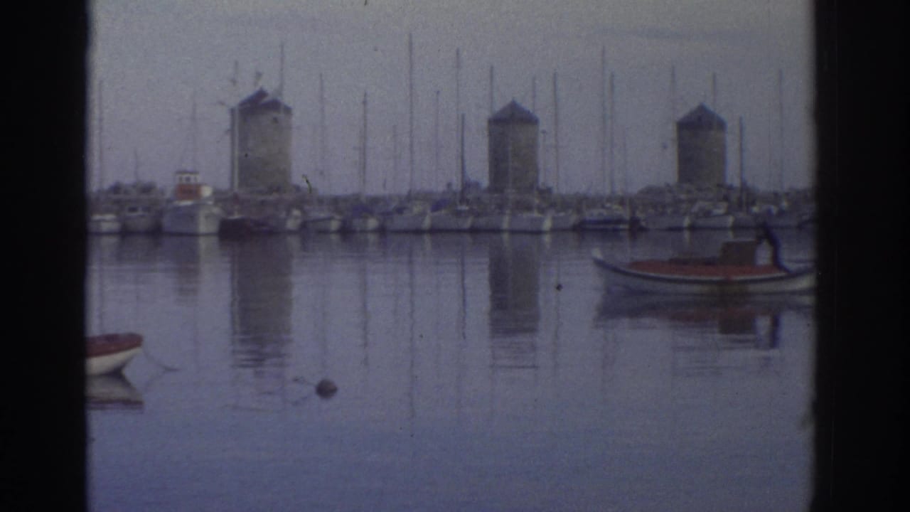 Stone windmill towers behind sailboat masts, small red-hulled boat in foreground water, 8mm.