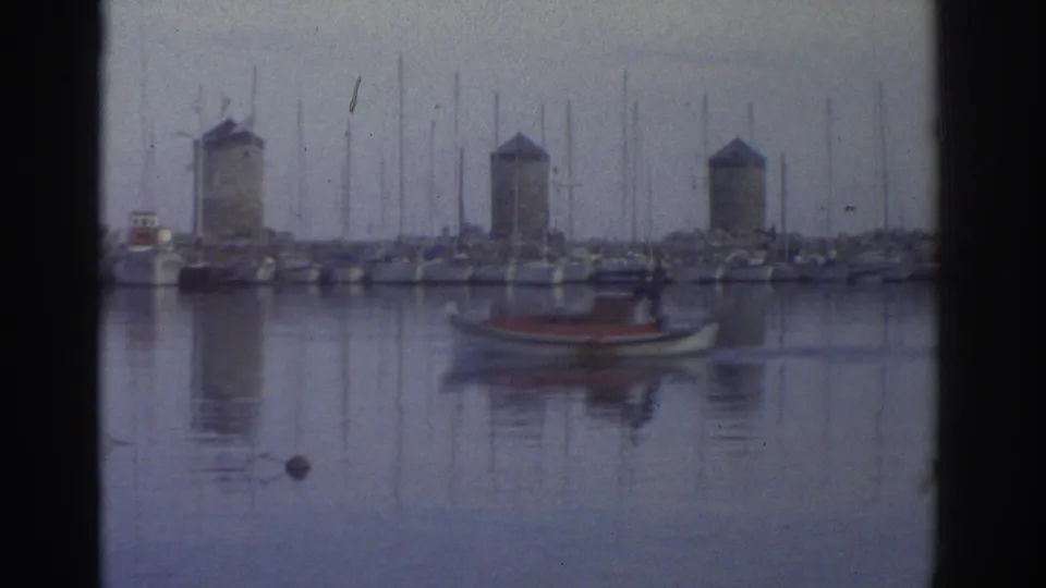 Wide shot of Greek harbor circa 1976 showing three stone windmill towers above a crowded marina, with a small red boat in the foreground and calm reflective water.