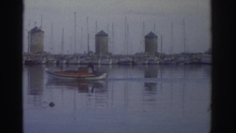 Greek harbor circa 1976 with stone windmill towers above packed marina; small red boat in foreground, blue-lavender color cast from aged 8mm film stock.
