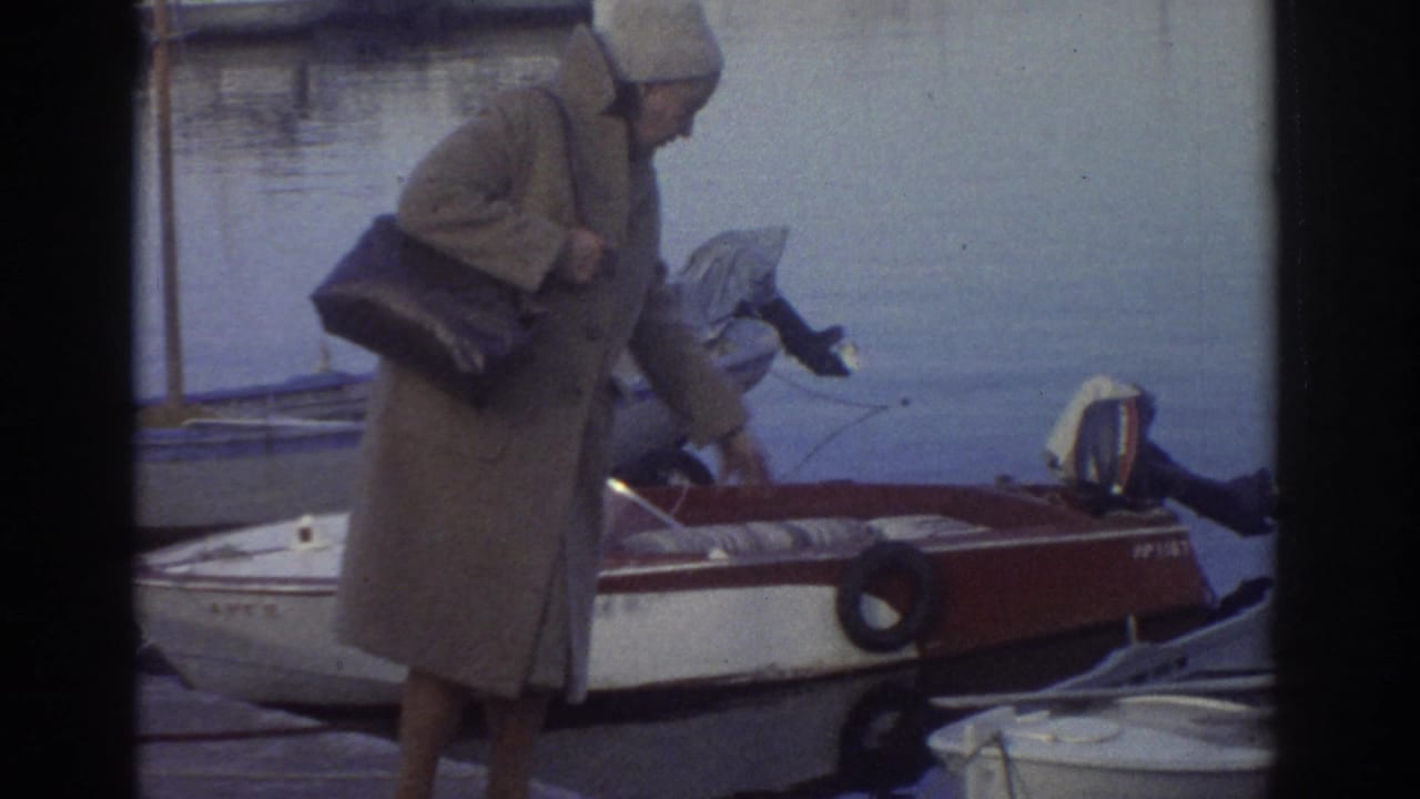 8mm home movie still, Greece 1976: figure in long tan coat with dark bag at harbor quayside next to red-and-white motorboat; second figure crouches alongside.