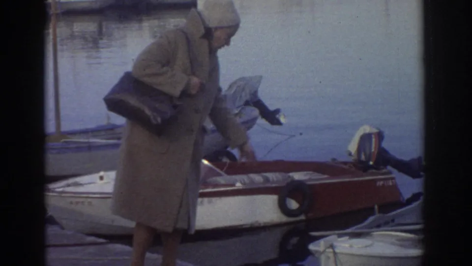 8mm home movie still, Greece 1976: figure in long tan coat with dark bag at harbor quayside next to red-and-white motorboat; second figure crouches alongside.