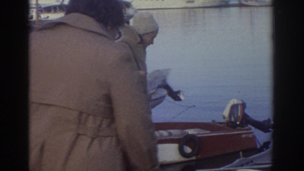 Close rear view of a person in a tan overcoat and light hat standing at a harbor; red boat hull and calm water visible to the right.