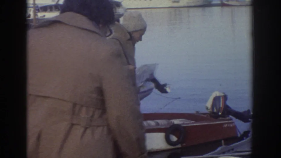 8mm home movie still, Greece 1976: close rear view of figure in tan overcoat and light hat at harbourside; red boat hull and calm water to the right.
