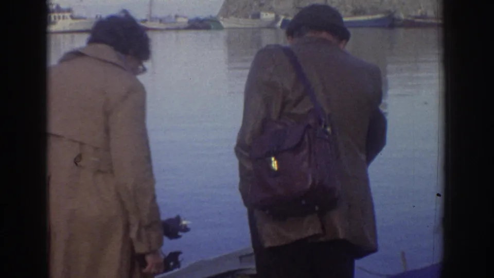 8mm home movie still, Greece 1976: two figures in coats viewed from behind at harbor quayside; distant stone fortification and harbor buildings visible across calm water.