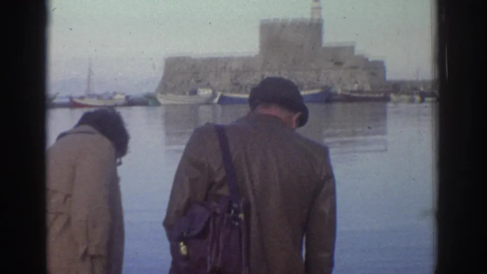 8mm home movie still, Greece 1976: two figures in coats at harbor quayside; a substantial stone fortress tower — consistent with Greek medieval harbor defenses — dominates the far shore.
