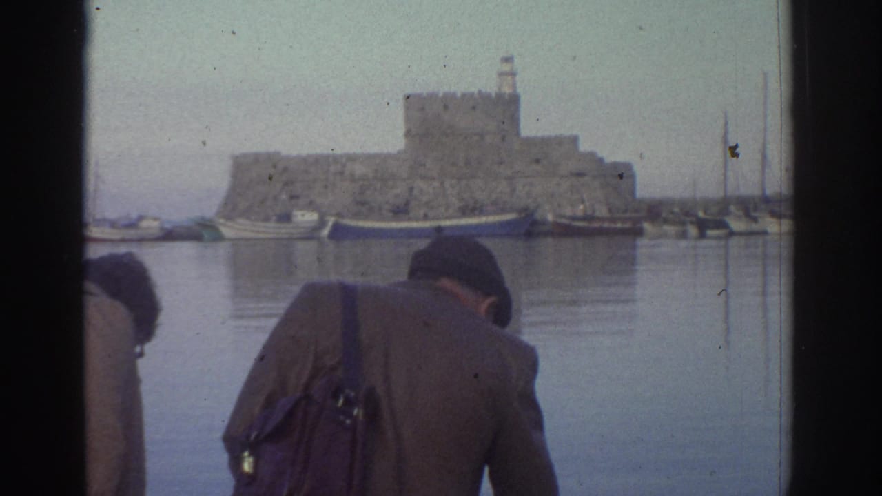 Two tourists at a Greek harbor in 1976, seen from behind, looking across calm water toward a medieval stone fortress with a tower.