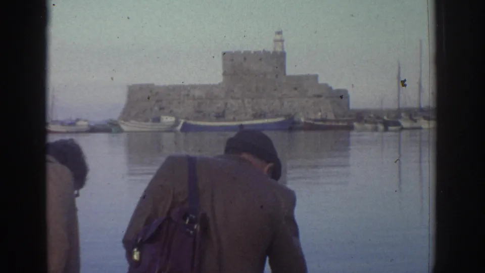 Two tourists at a Greek harbor in 1976, seen from behind, looking across calm water toward a medieval stone fortress with a tower.