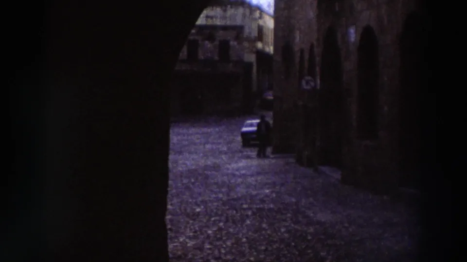 Medieval cobblestone street in a Greek old town, 1976, with stone arcaded buildings and a period-appropriate dark-colored automobile.