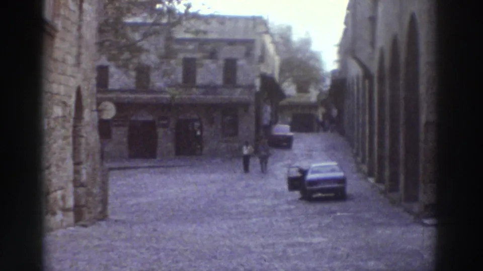 Open cobblestone square in a Greek old town, 1976, with medieval stone arcades, storefronts, a parked 1970s automobile, and scattered distant pedestrians.