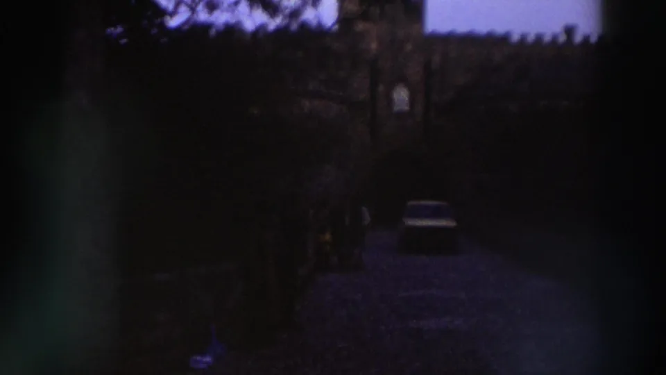 8mm home movie c.1976, Greece — light sedan near a crenellated stone gateway, deep purple-blue dye shift, dark foreground vignetting.