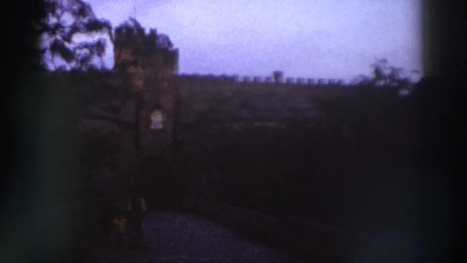 8mm frame c.1976, Greece — crenellated stone wall and tower, arched gateway opening, small figure near the base, trees on the left.
