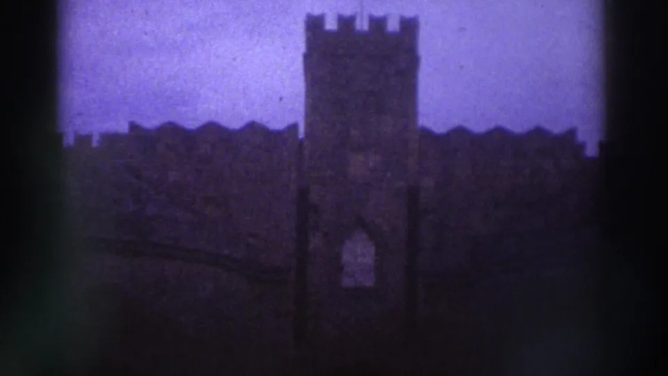 8mm frame c.1976, Greece — a square crenellated stone tower dominates the frame against a purple-blue aged-film sky; battlemented wall extends right.