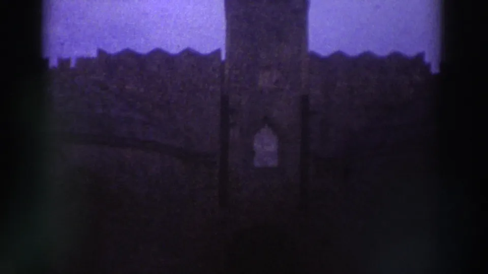 Underexposed 8mm frame with strong purple-blue color cast showing crenellated exterior walls of what appears to be a medieval stone fortification, Greece circa 1976.