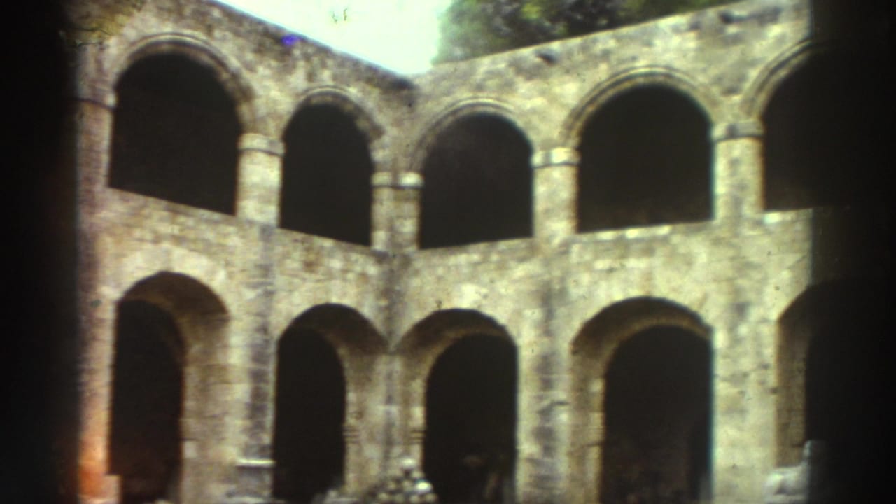 Brightly lit interior courtyard of a medieval stone structure in Greece circa 1976, showing two levels of Romanesque rounded arches in warm ochre stonework.