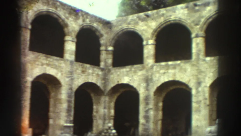 Brightly lit interior courtyard of a medieval stone structure in Greece circa 1976, showing two levels of Romanesque rounded arches in warm ochre stonework.