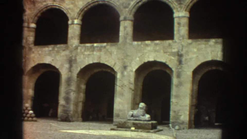 8mm home-movie frame of a medieval stone courtyard in Greece circa 1976: a sphinx statue on a plinth, a stack of cannonballs at lower left, and two tiers of Romanesque arches.