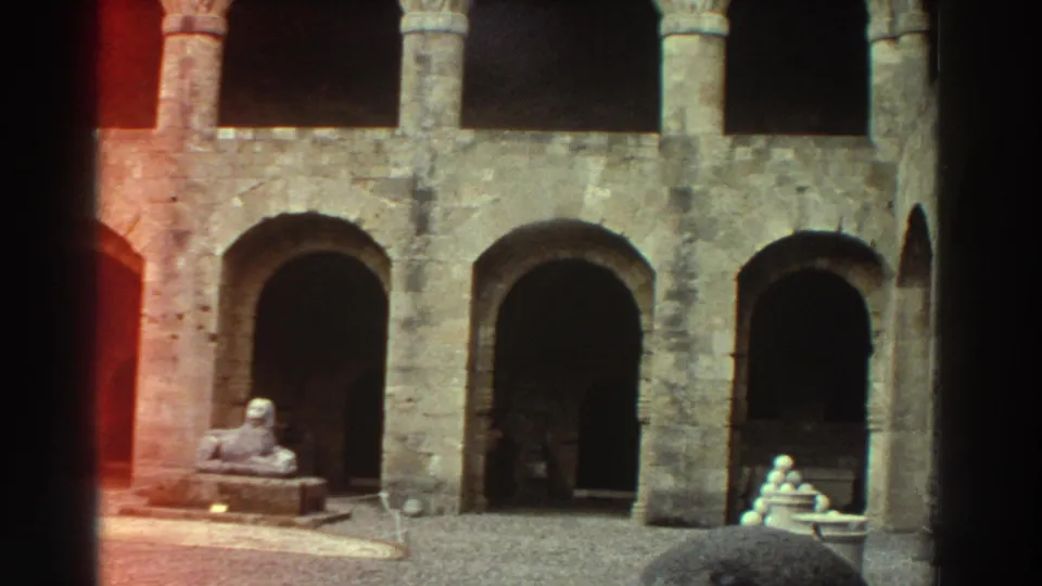 8mm home-movie frame of a Greek medieval courtyard circa 1976: sphinx statue, cannonball pyramid at right, central stone pillar, and a red film-burn streak entering from the left edge.