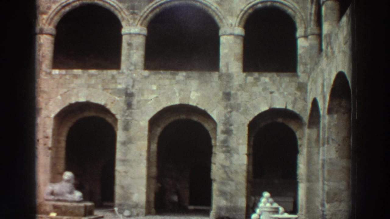 8mm film frame, Greece 1976 — two-tiered stone colonnade with rounded arches, a sphinx-like sculpture at lower left, and small figures near the staircase base; dim, interior-facing perspective.