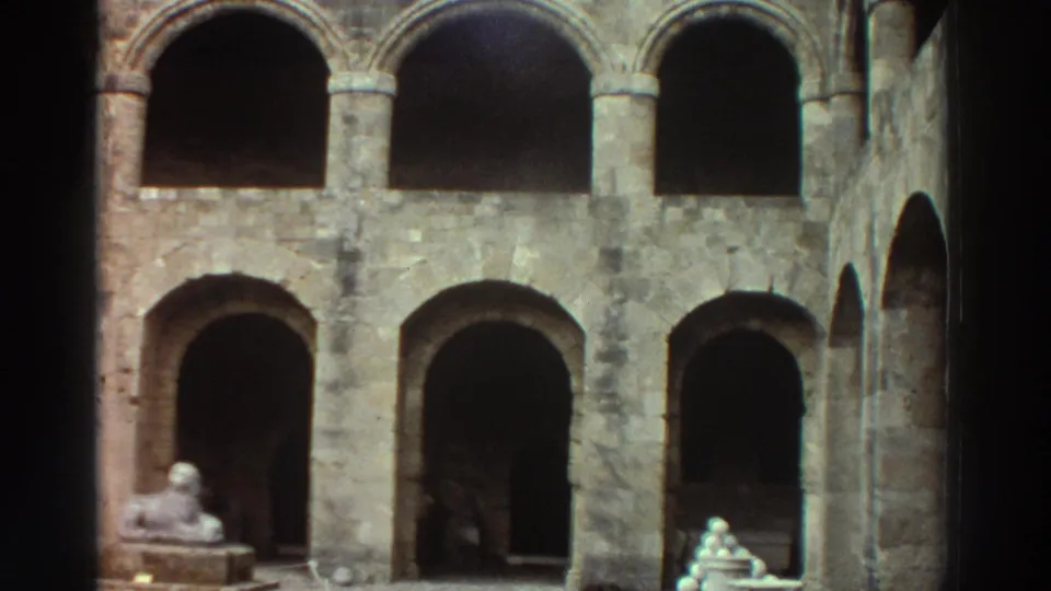 8mm film frame, Greece 1976 — two-tiered stone colonnade with rounded arches, a sphinx-like sculpture at lower left, and small figures near the staircase base; dim, interior-facing perspective.