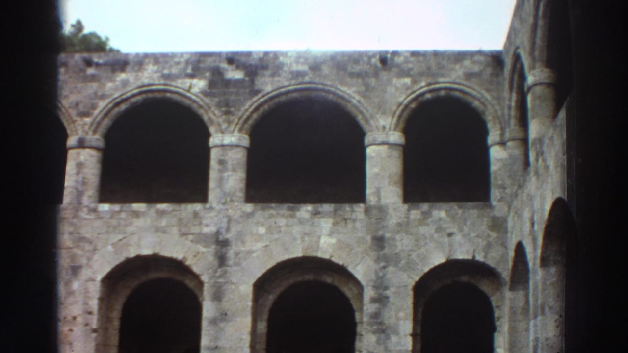8mm film frame, Greece 1976 — wide shot of two-tiered arched stone colonnade with open pale blue sky at upper right; no figures clearly discernible in this frame.