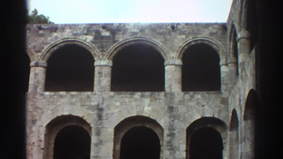 8mm film frame, Greece 1976 — wide shot of two-tiered arched stone colonnade with open pale blue sky at upper right; no figures clearly discernible in this frame.