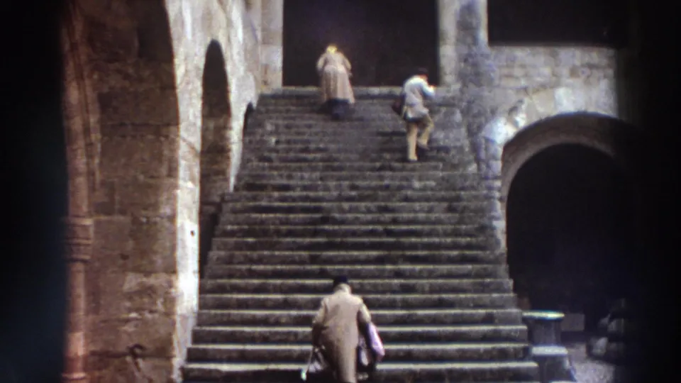8mm film frame, Greece 1976 — two visitors climbing a wide stone staircase past arched side openings; woman in light coat ascending, second figure in darker clothing behind her.