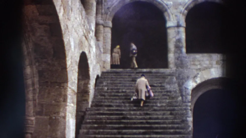 8mm film frame, Greece 1976 — visitors higher on the wide stone staircase; figure in beige coat with handbag at lower right, second figure visible within an arched passage at left.