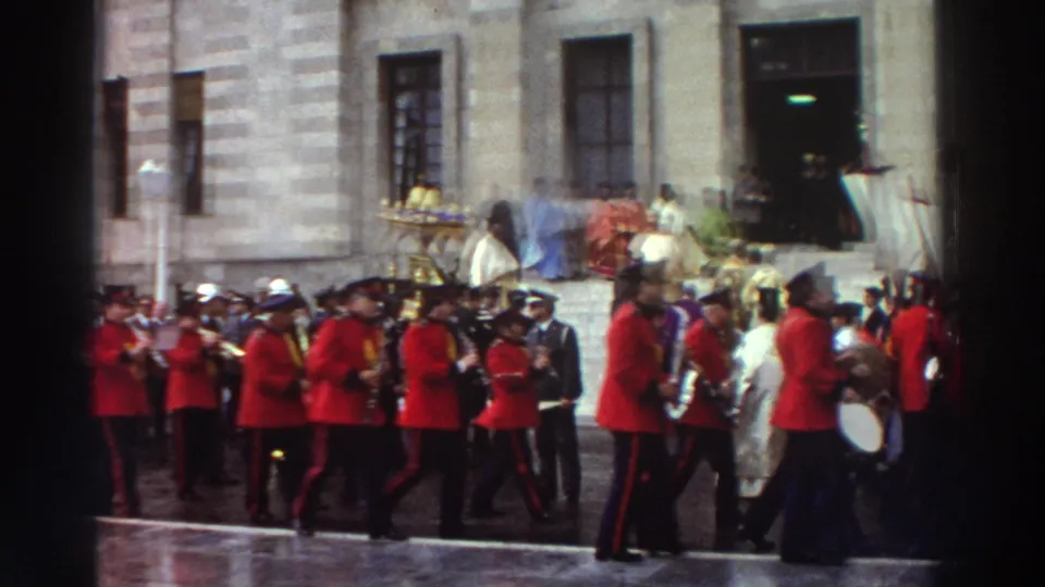 Full ceremonial procession across a wet stone courtyard in Greece, 1976; marching band in red, Orthodox clergy in white and gold, stone building facade with rectangular windows behind.