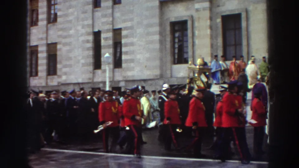 Continuing ceremonial procession in Greece, 1976; red-uniformed band, clergy in purple, white, and gold robes, and gathered crowd on wet stone paving before a stone building.