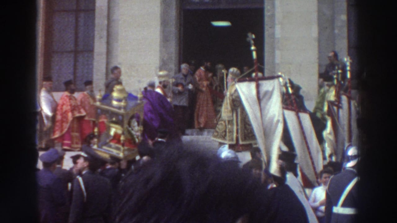 Clergy in red and gold vestments carry a large gilded religious icon near a church doorway, surrounded by a dense crowd.
