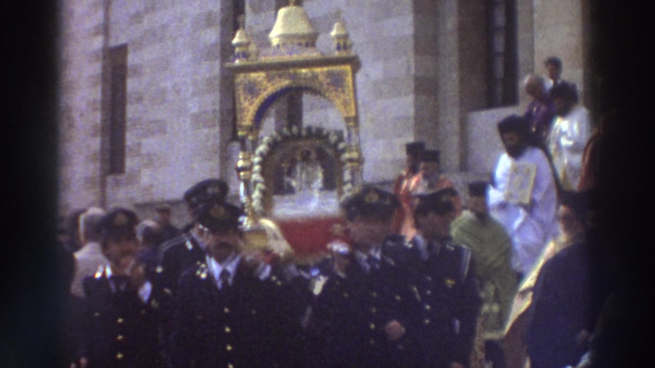 1976 Greece home-movie frame showing dark-uniformed men walking before an ornate gold religious reliquary near a stone church exterior.