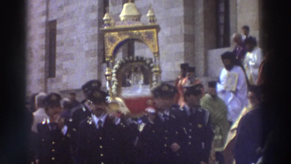 1976 Greece home-movie frame showing dark-uniformed men walking before an ornate gold religious reliquary near a stone church exterior.