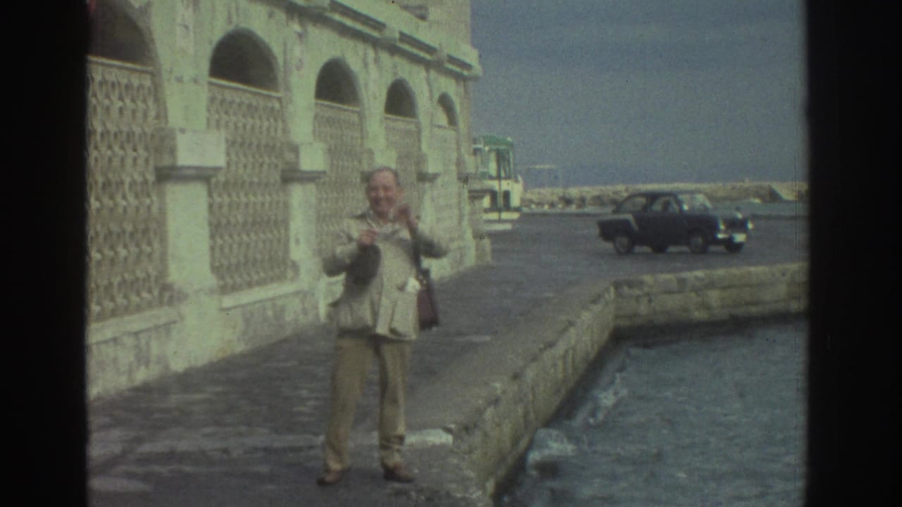 1976 Greece 8mm frame showing a man in a light jacket on a seaside stone pier with a dark sedan and arched waterfront building nearby.