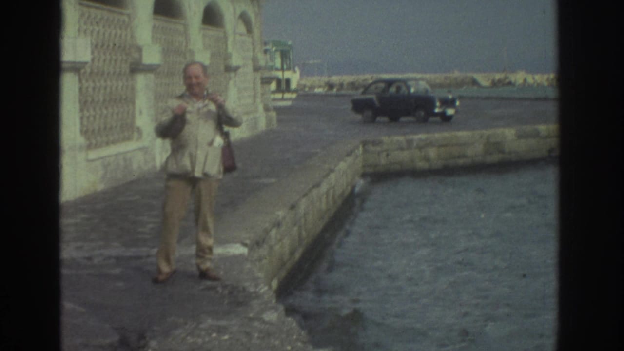 1976 Greece travel frame with a man smiling toward the camera on a stone waterfront, sea at right, and a dark sedan in the background.