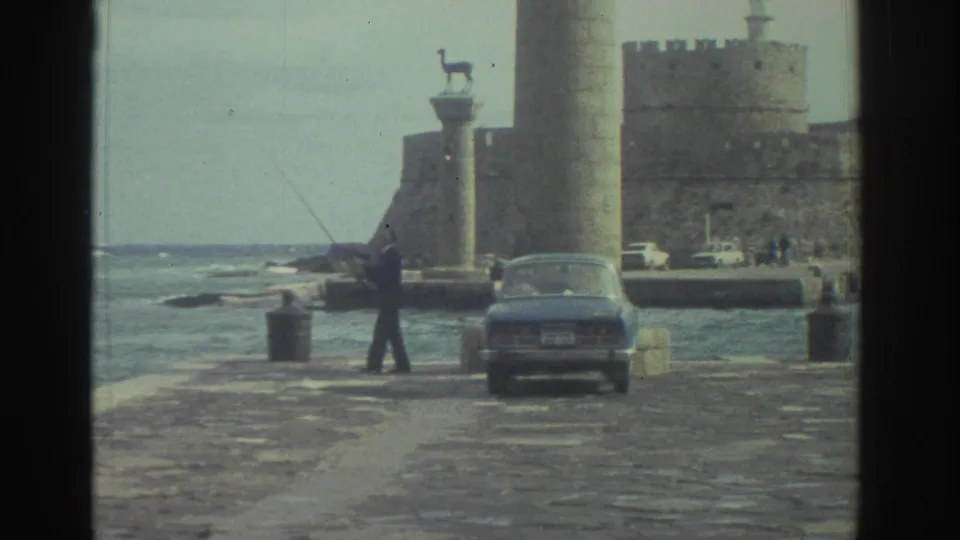 1976 Greece 8mm frame showing a fisherman casting near choppy water, with a parked sedan, tall column, animal statue, and castle-like wall behind.
