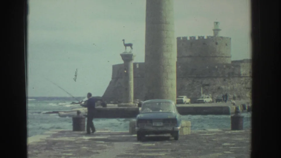 1976 Greece home-movie frame of a fisherman on a windy waterfront, with a sedan, column, animal statue, and fortified stone wall in view.