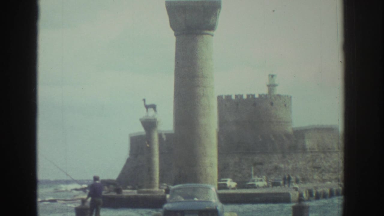 1976 Greece home-movie frame showing Heraklion harbor columns, distant fort walls, parked cars, and small figures near the water.