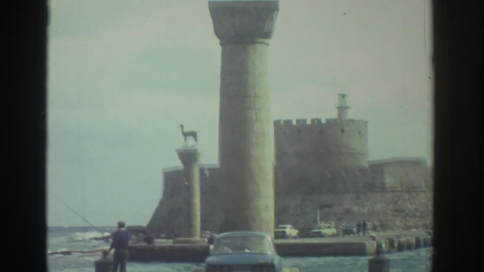 1976 Greece home-movie frame showing Heraklion harbor columns, distant fort walls, parked cars, and small figures near the water.