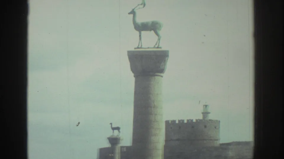 1976 Greece travel frame with a deer-topped harbor column in the foreground and fort walls behind it.