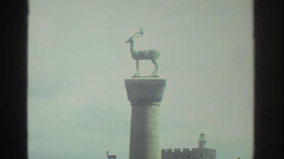 1976 Greece 8mm frame focused on a deer sculpture atop a harbor column, with fort masonry partly visible below.