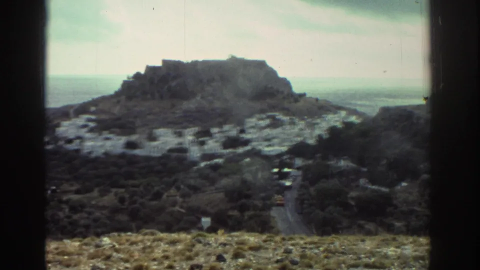 1976 Greece home-movie landscape showing a hillside settlement, winding road, rocky ground, and coastal water in the distance.