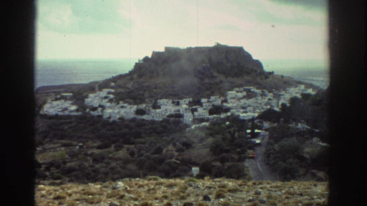 Wide view of white buildings below a rocky hilltop with sea beyond and dark vertical borders.