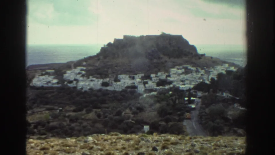 1976 Greece home-movie view of a coastal village below a rocky hilltop fortress, with sea in the distance and dark frame edges.