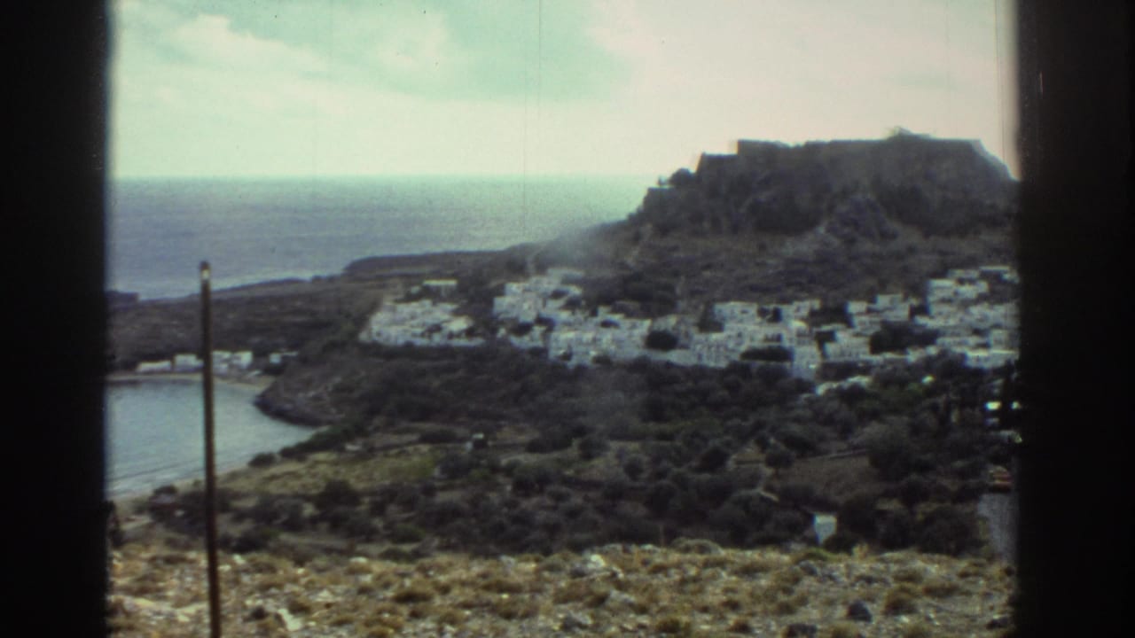 1976 Greece view of white buildings at the base of a rocky hill, with a curved inlet and open sea beyond.