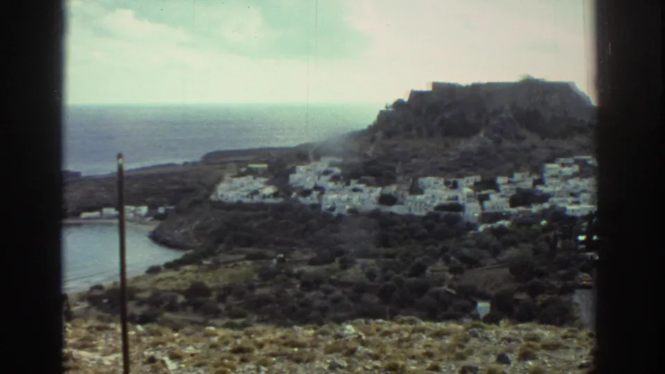 1976 Greece view of white buildings at the base of a rocky hill, with a curved inlet and open sea beyond.