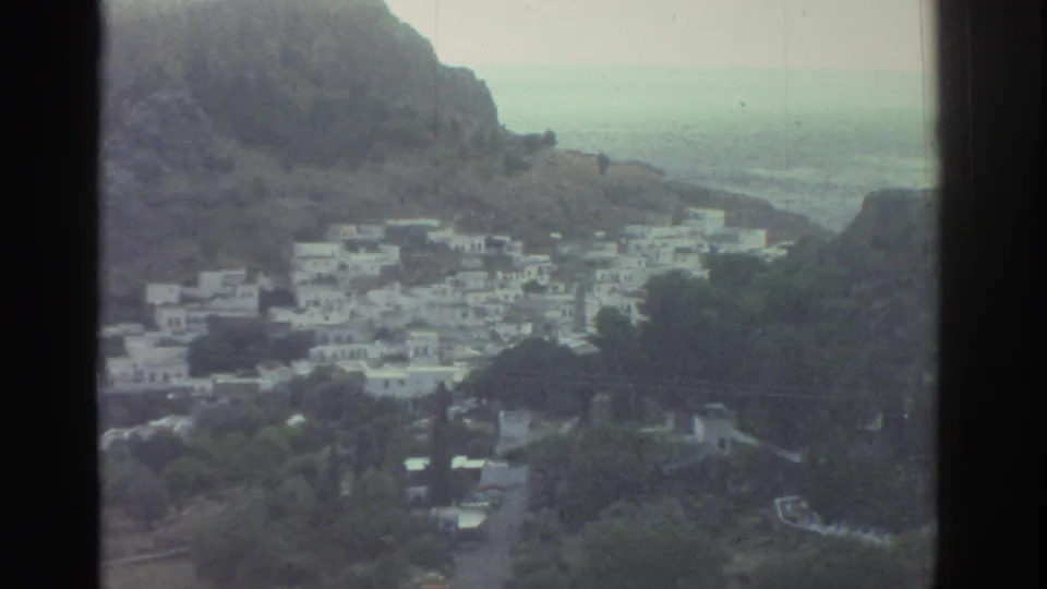 Closer 1976 Greece home-movie frame showing white village houses, trees, road access, rocky slope, and sea haze beyond.