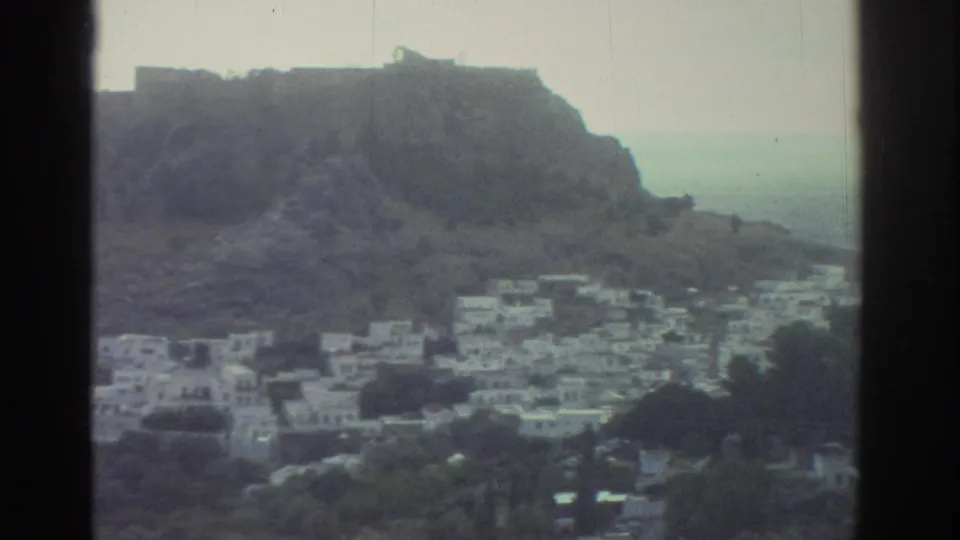 1976 Greece frame of clustered white buildings below a rocky hilltop fortress-like structure, with hazy sea in the background.