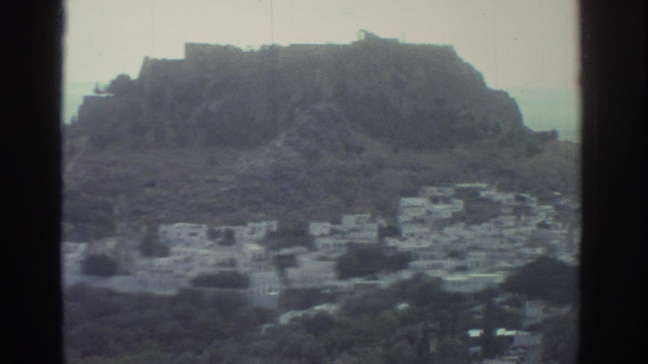 1976 Greece 8mm frame showing a whitewashed hillside village beneath a rocky ridge with the sea beyond.